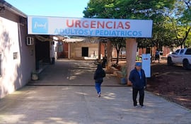 Mujer caminando hacia urgencias del hospital y hombre de pie con chaqueta y gorro en un ambiente bullicioso.