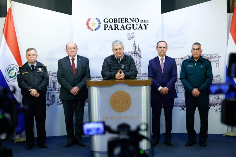 Cinco hombres en un estrado, uno en uniforme de policía, otro de cabello canoso en traje oscuro, y un portavoz en abrigo oscuro.