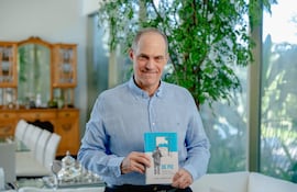 Jorge Mendelzon sonríe mientras sostiene su libro, vestido con una camisa azul en un entorno elegante y iluminado.