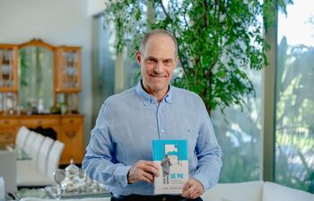 Jorge Mendelzon sonríe mientras sostiene su libro, vestido con una camisa azul en un entorno elegante y iluminado.