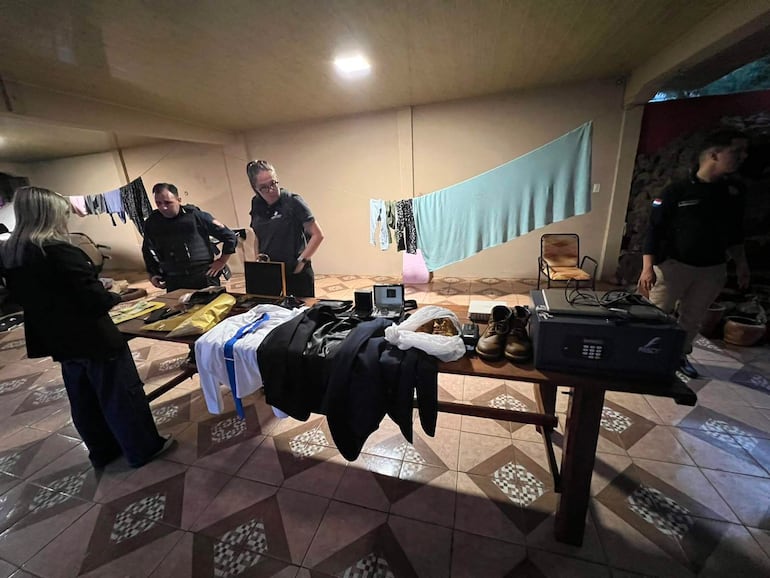 Tres hombres con chalecos oscuros revisan objetos sobre una mesa, mientras una mujer de chaqueta negra observa en el fondo sencillo.