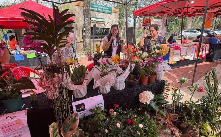 Mujer sosteniendo micrófono y otra con ramo de flores en un colorido stand de feria de flores al aire libre.