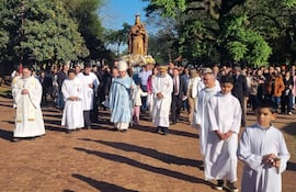 Multitud de personas y sacerdotes en vestimenta blanca en procesión religiosa bajo un cielo soleado rodeados de árboles.