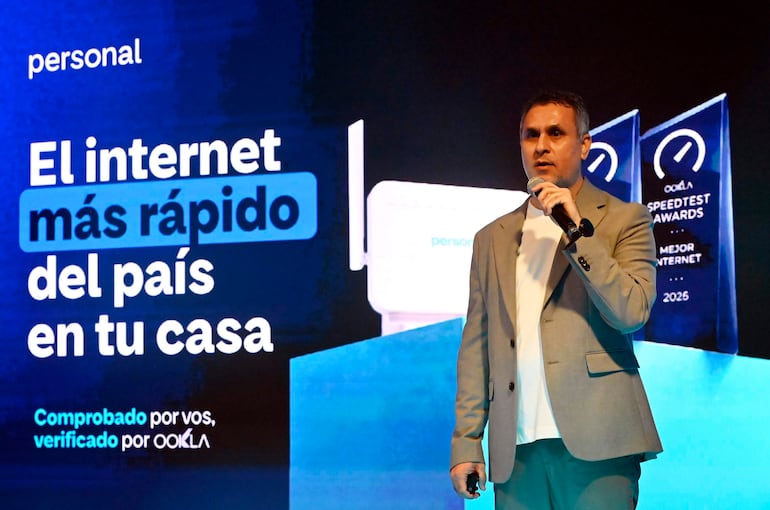Hombre en traje claro sostiene un micrófono, hablando ante un público, con carteles de 'internet más rápido' y trofeos visibles.