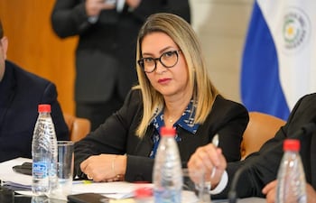 Mónica Recalde con gafas y expresión seria, sentada en una mesa con un hombre en perfil. Botellas de agua y aperitivos visibles.