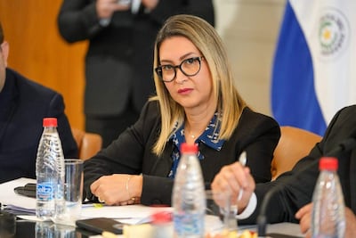 Mónica Recalde con gafas y expresión seria, sentada en una mesa con un hombre en perfil. Botellas de agua y aperitivos visibles.