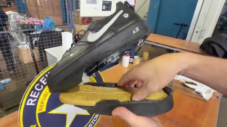 Un individuo sostiene una zapatilla negra de Nike mostrando su suela, mientras en el fondo se observan varias personas en un ambiente cerrado.