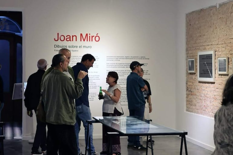Hombre con chaqueta verde gesticula, mientras dos hombres y una mujer conversan en una galería con obras de Joan Miró.