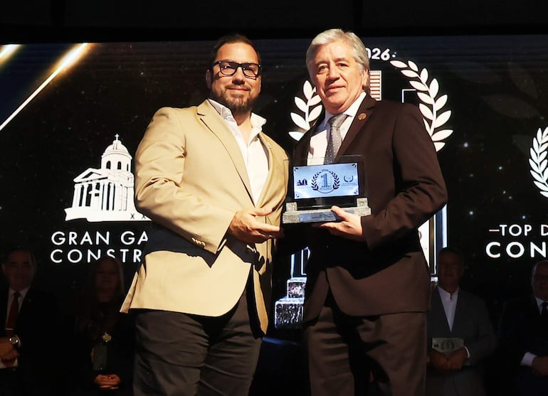 Hombre con saco claro y camisa blanca entrega un trofeo a hombre de saco oscuro, ambos sonrientes en un evento formal.