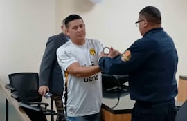 Hombre con camiseta blanca es esposado por un oficial en sala de audiencias.