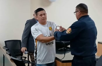 Hombre con camiseta blanca es esposado por un oficial en sala de audiencias.