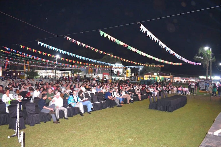Amplio público sentado en sillas dispuestas en filas, decoraciones festivas y escenario iluminado de la Fiesta Nacional de las Colectividades.