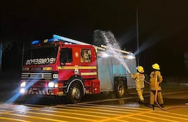 Bomberos con cascos amarillos luchan contra un incendio desde un camión rojo en área urbana iluminada por luces azules.