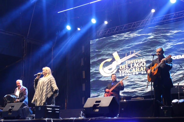 Mujer rubia con micrófono al centro del escenario, acompañada de músicos con guitarra y acordeón bajo luces nocturnas.