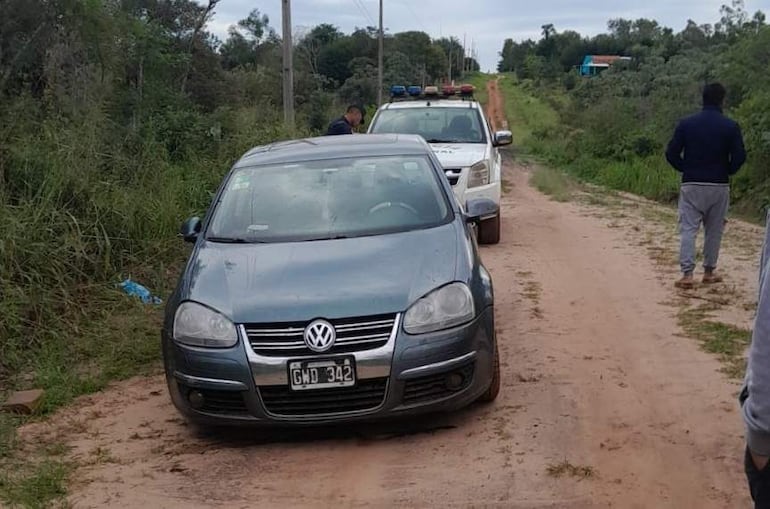Dos agentes policiales cerca de un Volkswagen gris oscuro, una persona en sudadera oscura observa la escena en un camino de tierra.