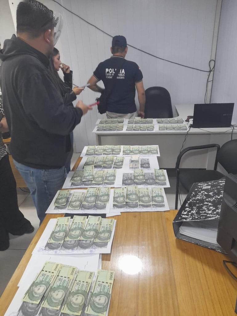 Oficiales de policía supervisan la situación mientras fajos de billetes y documentos están en la mesa.
