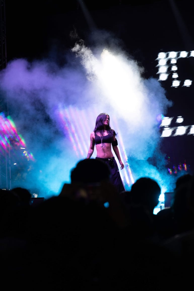 Artista femenina en escenario con luces de colores, vestida con top corto y falda larga, rodeada de una multitud entusiasta.