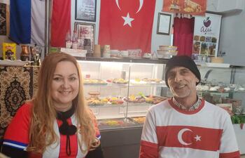Mujer con camiseta roja y hombre con camiseta blanca con bandera turca, sonriendo en un ambiente acogedor de cafetería decorada con productos turcos.