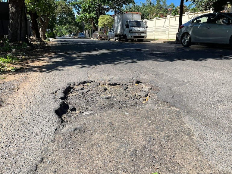 Bache visible en calle con camión blanco y automóvil plateado estacionados, iluminados por luz natural en un día soleado.