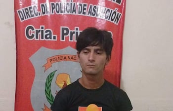 Joven de camiseta negra con diseño colorido frente a bandera de la Policía Nacional, postura erguida y expresión seria.