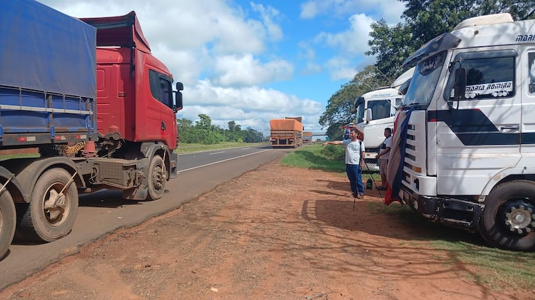 Tres hombres, uno con camiseta blanca y otro con bandera, conversan junto a camiones rojos y blancos en una carretera.