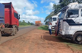 Tres hombres, uno con camiseta blanca y otro con bandera, conversan junto a camiones rojos y blancos en una carretera.