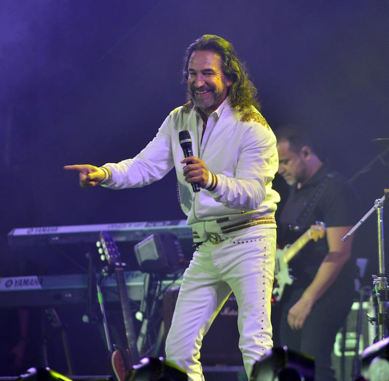 Marco Antonio Solís en traje blanco con detalles dorados, sosteniendo un micrófono y sonriente, interactuando con el público en un escenario iluminado.