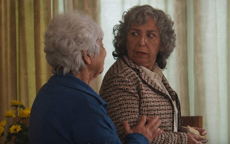 Amada Gómez con chaqueta azul gesticula mientras Olga Vallejos escucha atentamente en una sala decorada con flores amarillas.