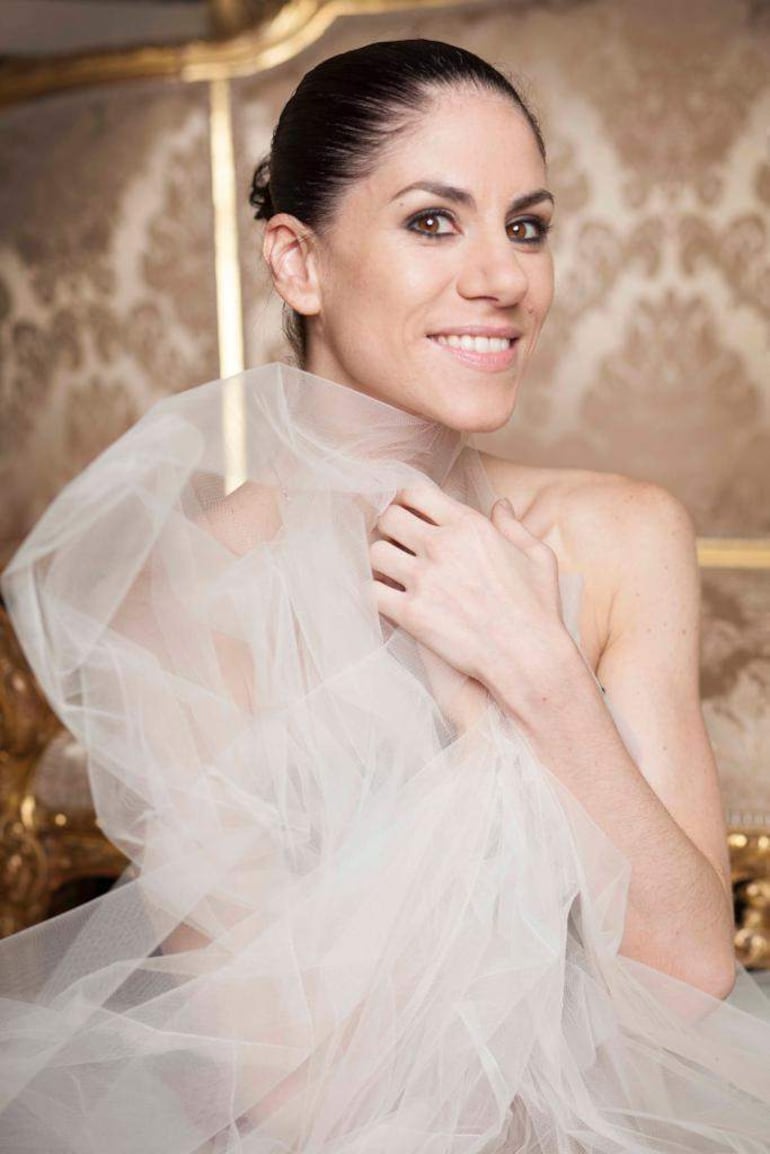 Mujer sonriente en vestido translúcido con peinado recogido, posando en un entorno elegante y decorativo.
