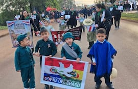 Cuatro niños en camisetas verdes y un poncho azul sostienen un cartel con banderas en una calle con árboles y carpas.