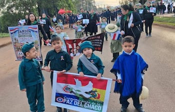 Cuatro niños en camisetas verdes y un poncho azul sostienen un cartel con banderas en una calle con árboles y carpas.