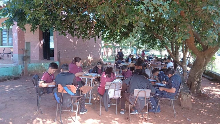 Grupo de estudiantes en uniformes burdeos y negro, sentados bajo un árbol, atentos a una docente en el centro.