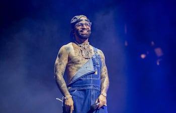 Artista robusto en overoles de mezclilla y camiseta, luciendo varios tatuajes y gafas, señalando a la audiencia en un ambiente festivo.