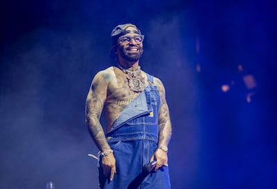 Artista robusto en overoles de mezclilla y camiseta, luciendo varios tatuajes y gafas, señalando a la audiencia en un ambiente festivo.