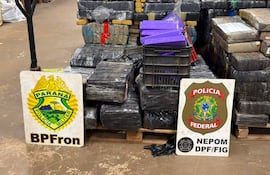 Paquetes envueltos en plástico negro apilados en bodega, con carteles de la Policía Militar de Paraná y Policía Federal de Brasil.