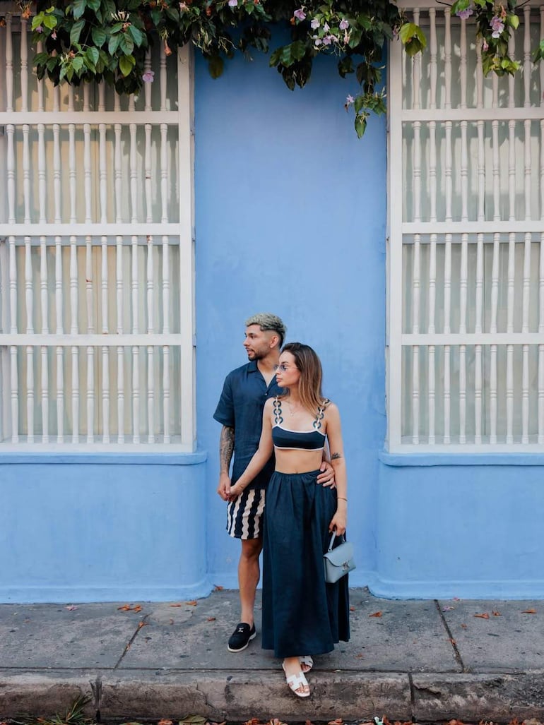Julio Enciso, con camisa azul y pantalones a rayas, sostiene de la mano a Melissa Cardona, en bikini azul y falda larga oscura, junto a una pared azul.