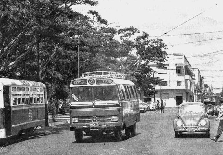 Dos autobuses en la calle; uno grande con '32' y otro rojo y blanco. Ambiente soleado con árboles al fondo.