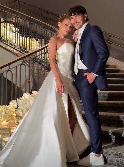 Pareja sonriente en escalinata, ella con vestido de novia blanco y él en traje azul oscuro, rodeados de flores y velas.