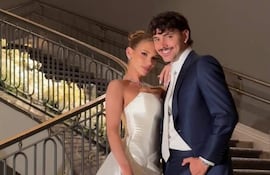 Pareja sonriente en escalinata, ella con vestido de novia blanco y él en traje azul oscuro, rodeados de flores y velas.