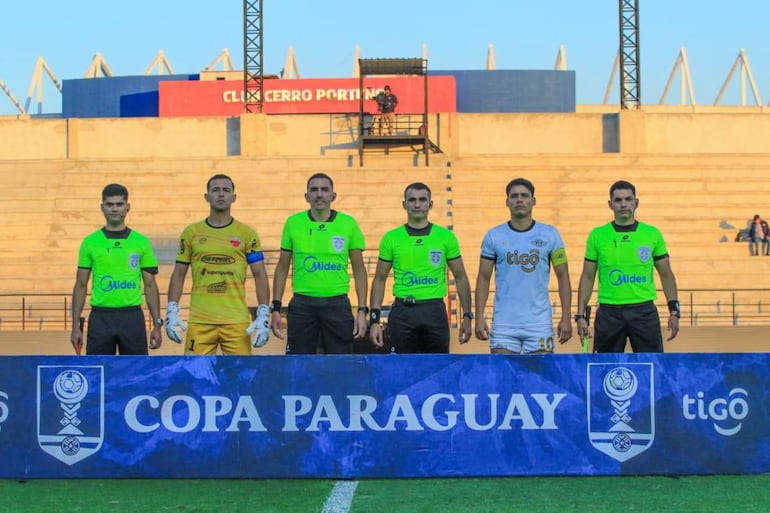 Grupo de seis personas en el estadio, árbitros en camisetas verdes y jugadores en amarillas y blancas, ambiente de preparación.