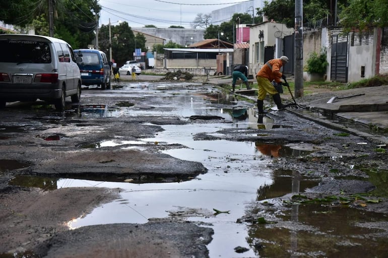 Tres trabajadores en overoles naranja y verde limpian la calle con charcos, en un ambiente urbano dañado por la lluvia.