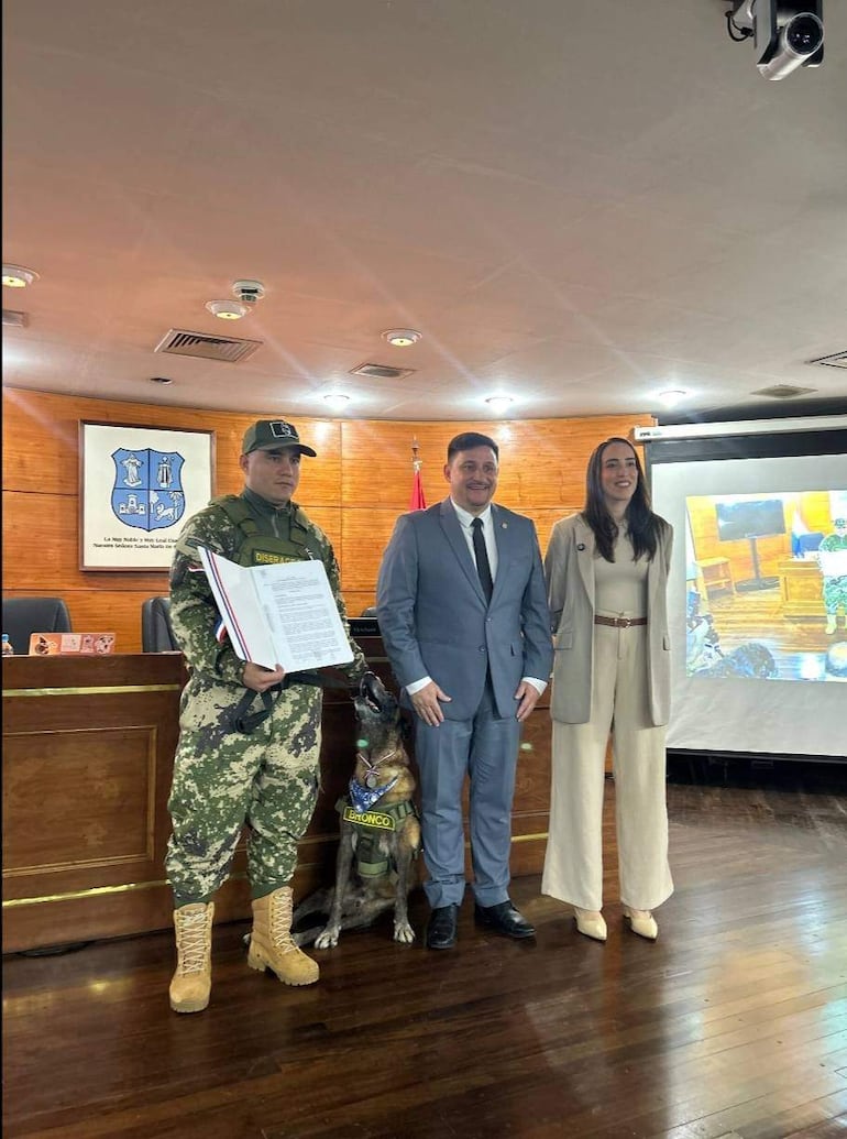 Sgto Gustavo Gómez con uniforme militar y documento, junto a un hombre en traje gris y una mujer sonriente en sala de conferencias.