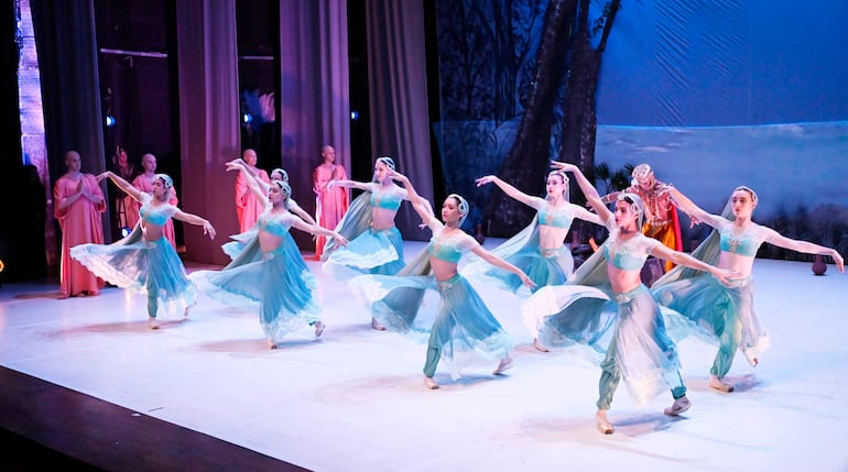 Ocho bailarinas en trajes celestes y plateados realizan una coreografía en un escenario iluminado, con figuras secundarias de fondo.