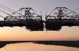 Puente colgante en construcción, cables tensores y andamiaje, con un río y vegetación al fondo bajo colores de atardecer.