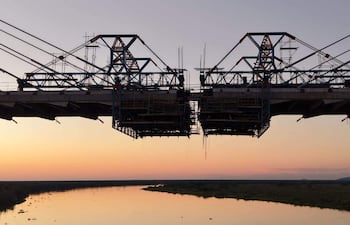 Puente colgante en construcción, cables tensores y andamiaje, con un río y vegetación al fondo bajo colores de atardecer.