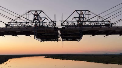 Puente colgante en construcción, cables tensores y andamiaje, con un río y vegetación al fondo bajo colores de atardecer.