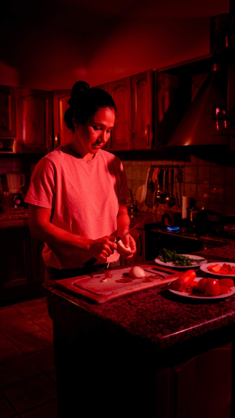 Mujer con cabello recogido y camiseta clara corta cebollas en un ambiente de cocina con luz tenue y roja.