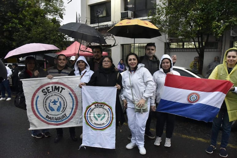 Mujer con abrigo blanco sostiene bandera paraguaya y sonríe; otras con carteles y paraguas en un ambiente urbano lluvioso.
