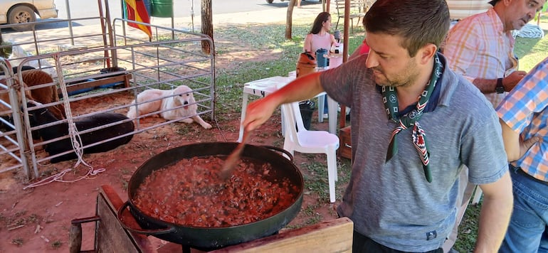 Rômulo Moraes en camiseta gris y pañuelo verde cocina un guiso, apoyado en un gran recipiente con fondo de personas y animales.