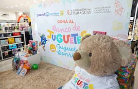 Un gran oso de peluche con camiseta del Banco de Juguetes rodeado de juguetes variados; ambiente de recolección en un local comercial.